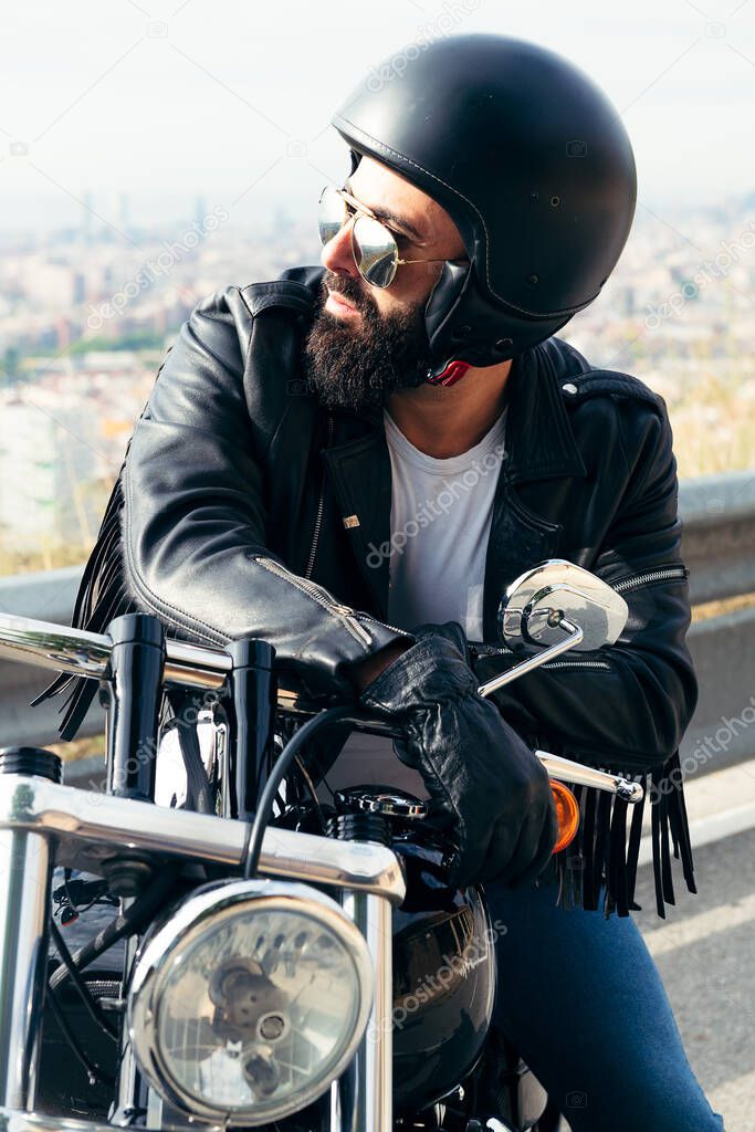 biker with helmet and leather jacket sitting on his motorbike with the city far away in the background, concept of freedom and rebel lifestyle