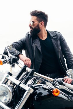 Deri ceketli, güneş gözlüklü, motosikletine yaslanmış, özgürlük ve motorcu yaşam tarzı kavramına sahip bir adamın dikey fotoğrafı.