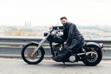 Deri elbiseli ve güneş gözlüklü bir adam yolda motosikletinin üzerinde oturuyor. Şehrin panoramik manzarası, özgürlük konsepti ve motorcu yaşam tarzı, metin için kopyalama alanı.
