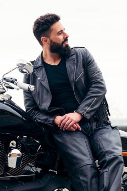 Deri giyinmiş yakışıklı sakallı bir adamın dikey fotoğrafı motosikletinin üstünde duruyor, özgürlük kavramı ve motorcu yaşam tarzı.