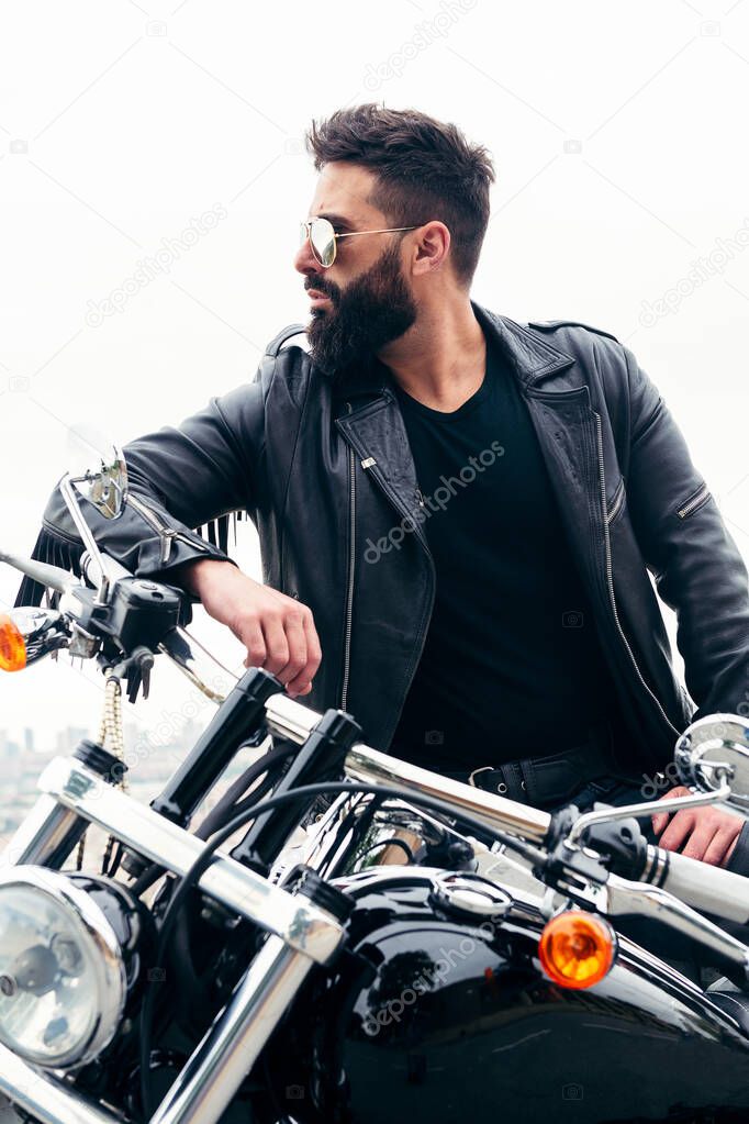 vertical photo of a man with leather jacket and wearing sunglasses leaning at his motorcycle, concept of freedom and biker lifestyle, copy space for text