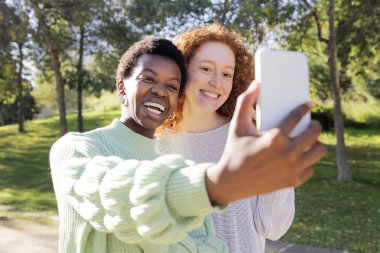 Mutlu gülümseyen ve parkta cep telefonuyla fotoğraf çeken iki genç kadın. Teknoloji ve arkadaşların eğlenmesi kavramı.