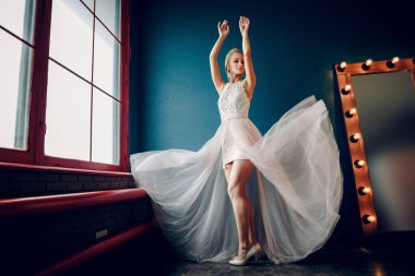 Havadar elbiseli, gözleri kapalı dans eden ellerini kaldıran bir sarışın. Elbisesi dalgalanıyor. Arka planda mavi bir duvar ve bir ayna var. Yan kırmızı pencere