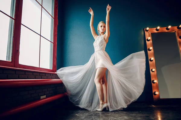 穿着轻飘飘的衣服的金发新娘正在跳舞 她的衣服在飘扬 背景是一堵深蓝色的墙和一面镜子 旁边是一个红色的窗户 — 图库照片