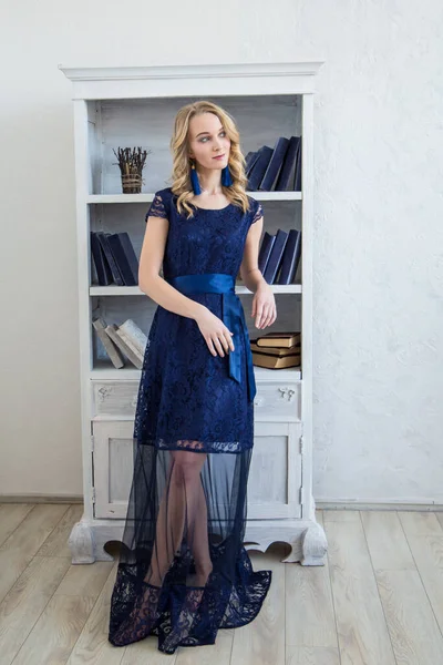 A tall and curly blonde with a slight smile in a blue dress stands in front of a bookcase with blue books