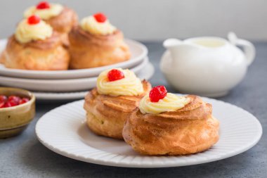 İtalyan pastası - zeppole di San Giuseppe, yakın çekim. Choux pastasından yapılmış kremalı börekler, krema ve vişne ile doldurulmuş. Aziz Yusuf Günü 'nü kutlamak için yenir. Gri masada beyaz tabak.