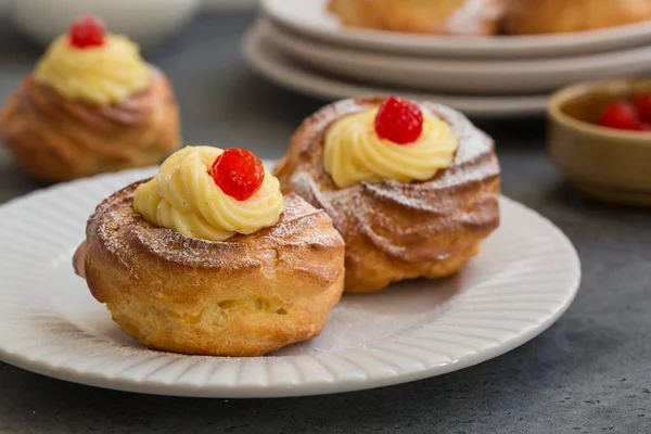 İtalyan pastası - zeppole di San Giuseppe, yakın çekim. Choux pastasından yapılmış kremalı börekler, krema ve vişne ile doldurulmuş. Aziz Yusuf Günü 'nü kutlamak için yenir. Gri arkaplanda beyaz plaka.