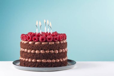 Beyaz masa ve mavi arka planda ahududu ve mumlarla çikolatalı doğum günü pastası. Boşluğu kopyala.