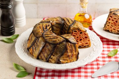 Patlıcan Timballo di Melanzane. Makarna ve domatesin etrafına ızgara patlıcan, peynir sosu ve fırın. İtalyan yemeği. Kırmızı ve beyaz masa örtülü bir masa..