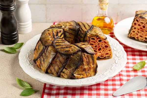 Patlıcan Timballo di Melanzane. Makarna ve domatesin etrafına ızgara patlıcan, peynir sosu ve fırın. İtalyan yemeği. Kırmızı ve beyaz masa örtülü bir masa..