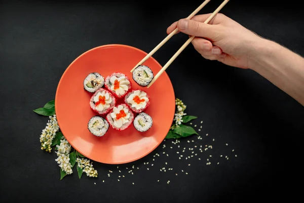 Kollarında suşi çubukları tutan bir adamın üst görüntüsü. Suşi ruloları siyah bir arka planda çiçekli tabağa konur..