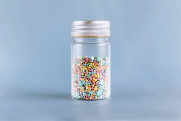 Renkli şekerleme serpiştirilmiş küçük cam kavanoz, mavi arka planda pasta ve fırın süslemesi.