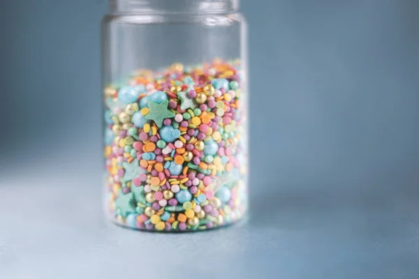 Renkli şekerleme serpiştirilmiş cam kavanoz, mavi arka planda pasta ve fırın süslemesi.