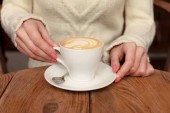 Zavřete ženské ruce s šálkem kávy cappuccino s pěnou s pěknou maskou. Perfektní červený gel na nehty. Dřevěný přirozený stůl. Zpracování tvůrčího barevného příspěvku