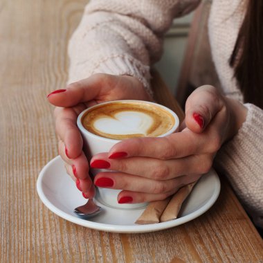 Fincan kahve kapuçino desen kalp ile köpük ile tutarak yakın çekim eller. Mükemmel kırmızı jel Lehçe manikür. Ahşap doğal masa. Yaratıcı renk sıcak mesaj işleme instagram tarzı.