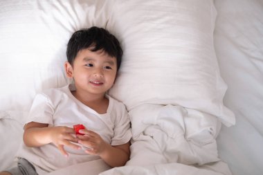 3 yıl eski küçük şirin Asyalı çocuk beyaz gömlek evde yatakta, çocuk oyun yalan ve beyaz yatak yastık ve battaniye ile gülümseyen, kopya alanı olan Üstten Görünüm Ekle metin.