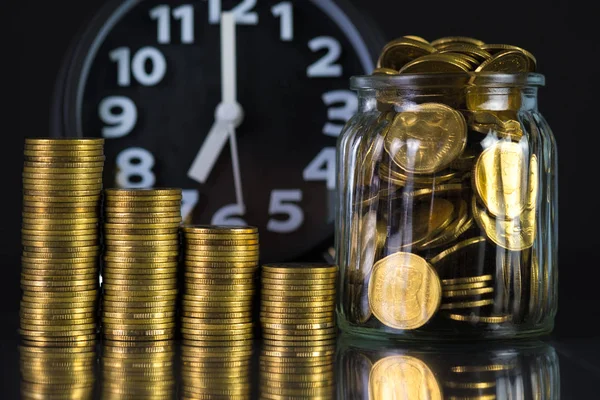 Coins stacks with coin in glass jar bottle and alarm clock in dark room, business and finance concept idea.