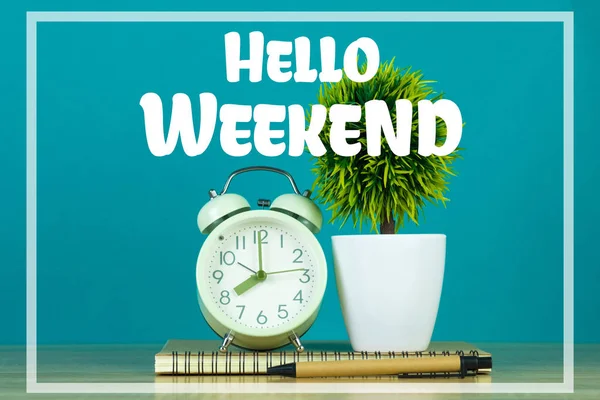 Hello Weekend text on green background and little tree in white vase with vintage alarm clock and notebook paper pen on wooden table.