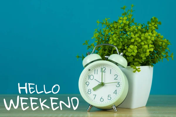 Hello Weekend text on green background and little tree in white vase with vintage alarm clock on wooden table.