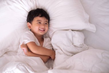 3 yıl eski küçük şirin Asyalı çocuk beyaz gömlek evde yatakta, çocuk oyun yalan ve beyaz yatak yastık ve battaniye ile gülümseyen, kopya alanı olan Üstten Görünüm Ekle metin.