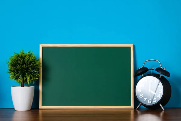 Blank green chalkboard with wood frame and little decorative tree in white vase and vintage alarm clock on wooden table with copy space for add text.