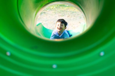 Küçük Asyalı bir çocuk yazın güneş ışığı altında oyun parkında kayağa tırmanıyor çocuklar okul bahçesinde oynuyor. Anaokulundaki mutlu çocuk.
