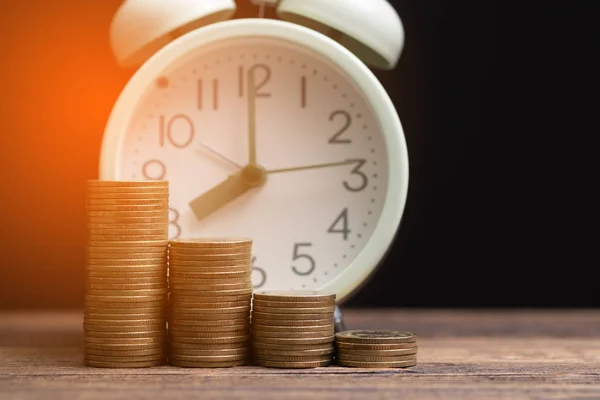 Reloj despertador y pilas de monedas en la mesa de trabajo en habitación oscura, tiempo —  Fotos de Stock