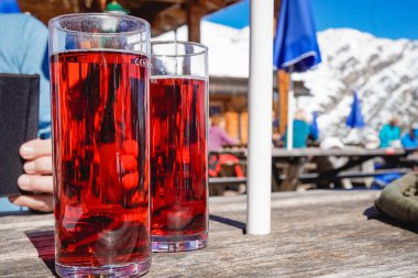 İki bardak kırmızı meyve suyu, güneşli gökyüzü olan dağlardaki bir restoranın dışındaki ahşap masada duruyor.