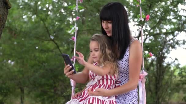 自然の中で電話を見て微笑む美しい若い母と娘 家族と技術の概念 — ストック動画