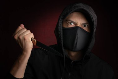 Yüz maskesi ve siyah beyzbol topu takan kızgın görünümlü bir hırsızı bıçakla tehdit ediyor.