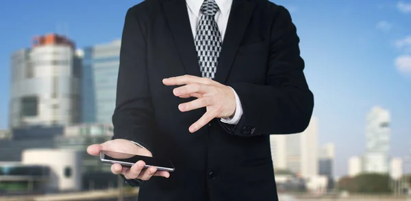 Businessman Holding Smartphone in Hand With Hand Gesture Over The Phones Screen With Business City and Corporate Buildings In Background — Stock Photo, Image