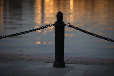 İşlenmiş demir bir çit. Gün batımında iskelede zincire vurulmuş bir sırık. Kaldırım kenarında hafif bir sis içinde dekoratif bir bariyer.
