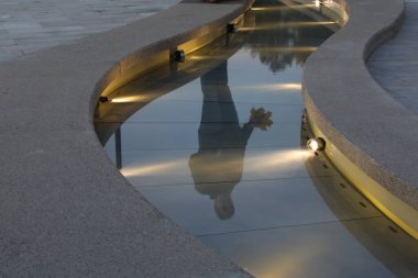 Taştan bir kanalda ışık saçan kıvrımlı bir akarsu. Bir insanın siluetinin yansıması. Akşam vakti şehrin eğlence parkı tasarımının dekoratif bir unsuru..