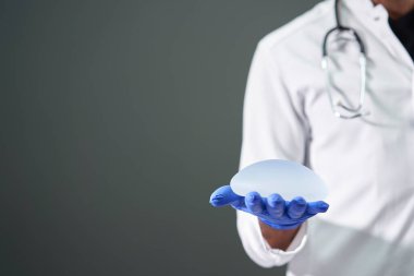 Bir doktorun ellerine silikon göğüs implantı. Beyaz önlüklü doktor, arka planda göğüs implantı olduğunu gösteriyor..