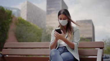 Şehirde çalışan bir kadın, bankta oturuyor, akıllı telefon kullanıyor. Koronavirüs salgını için maske takıyor. Mavi ceket ve kot pantolon giyiyor. Arkasında finans merkezinin bazı binaları var..