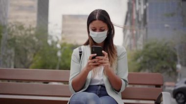 Şehirde çalışan bir kadın, bankta oturuyor, akıllı telefon kullanıyor. Koronavirüs salgını için maske takıyor. Mavi ceket ve kot pantolon giyiyor. Arkasında finans merkezinin bazı binaları var..