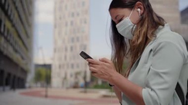Yüz maskesi takan, kulaklıklı akıllı telefon kullanan bir kadının yakın çekimi. Koronavirüs salgını için maske takıyor. O bir tarafta ve arka planda bazı binalar var. Mavi ceket ve kot pantolon giyiyor..
