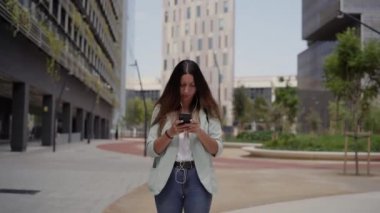 Çalışan bir kadının kulaklıkla arama yaptığı orta boy bir fotoğraf. Kameraya doğru yürüyor. Bir finans merkezinde, mavi ceket ve kot pantolon giyiyor..