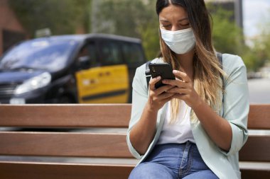Şehirde çalışan bir kadın, bankta oturuyor, akıllı telefon kullanıyor. Koronavirüs salgını için maske takıyor. Mavi ceket ve kot pantolon giyiyor. Arkasında finans merkezi ve tipik bir Barselona taksisi olan binalar var..