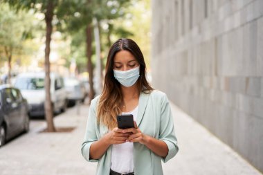 Şehirde çalışan bir kadının akıllı telefon kullanarak çekilmiş bir fotoğrafı. Koronavirüs salgını için maske takıyor. Kameraya bakıyor. Mavi bir ceket ve kot pantolon giyiyor..