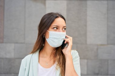 Çalışan bir kadının telefonda konuşurken çekilmiş yakın plan fotoğrafı. Kendisini koronavirüs salgını olarak gizliyor. Dümdüz, sağa bakıyor. Mavi bir ceket giyiyor. Arka plan gri bir duvar..