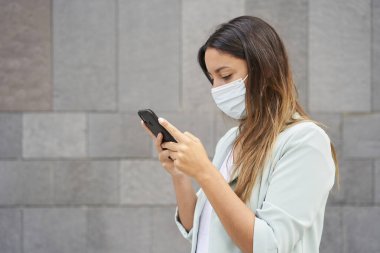 Akıllı telefonuyla maske yazan çalışan bir kadının orta fotoğrafı. Sol tarafta metni entegre etmek için boş bir alan var. Mavi ceket ve kot pantolon giyiyor. Arka plan gri bir duvar..