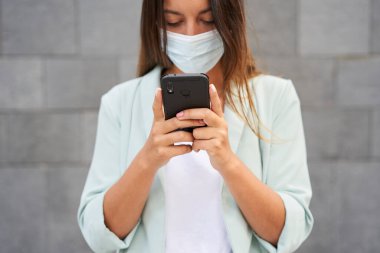 Şehirde çalışan bir kadının akıllı telefon kullanarak yakın plan çekimleri. Koronavirüs salgını için maske takıyor. Kameraya bakıyor. Mavi ceket ve kot pantolon giyiyor. Arka plan gri bir duvar..