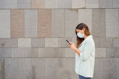 Yüz maskeli ve akıllı telefon kullanan bir kadının orta boy fotoğrafı. Sol tarafta metni entegre etmek için boş bir alan var. Mavi ceket ve kot pantolon giyiyor. Arka plan gri bir duvar..