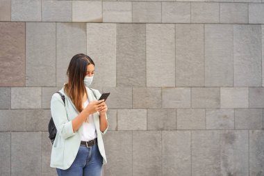 Akıllı telefonuyla maske takmış orta boy çalışan bir kadının fotoğrafı. Sağda metni entegre etmek için boş bir alan var. Mavi ceket ve kot pantolon giyiyor. Arka plan gri bir duvar..
