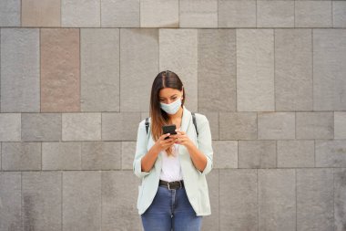Şehirde çalışan bir kadının akıllı telefon kullanarak orta boy görüntüsü. Koronavirüs salgını için maske takıyor. Kameraya bakıyor. Mavi ceket ve kot pantolon giyiyor..