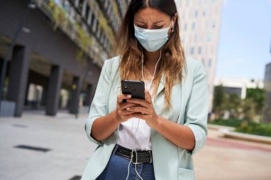 Finansal merkezde çalışan bir kadının akıllı telefon ve kulaklıkla çekilmiş bir fotoğrafı. Koronavirüs salgını için maske takıyor. Kameraya bakıyor. Mavi bir ceket ve kot pantolon giyiyor..