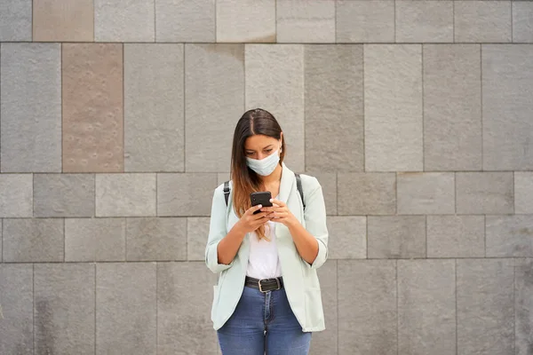 Şehirde çalışan bir kadının akıllı telefon kullanarak orta boy görüntüsü. Koronavirüs salgını için maske takıyor. Kameraya bakıyor. Mavi ceket ve kot pantolon giyiyor..