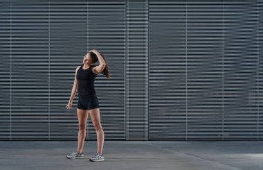 Beyaz bir kadın sokakta esneme egzersizleri yapıyor. Genç atlet kız gri bir metal duvarın önünde duruyor. Siyah spor giyer..