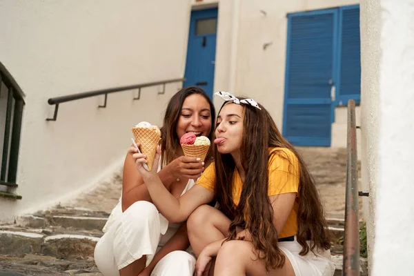 Young sisters eating ice cream and laughing sitting on some stairs in a pretty village. One gives the other ice cream. Concept of friendship, summer, family...
